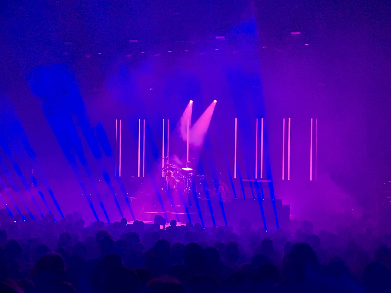 A view across a crowd of silhouetted people to a stage bathed in purple and pink lights where a performer stands in a pink spotlight.