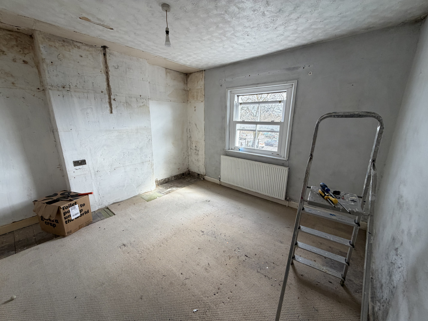 An empty room mid-renovation with stripped walls, stains, a window with a radiator below, and a step ladder on the carpet.