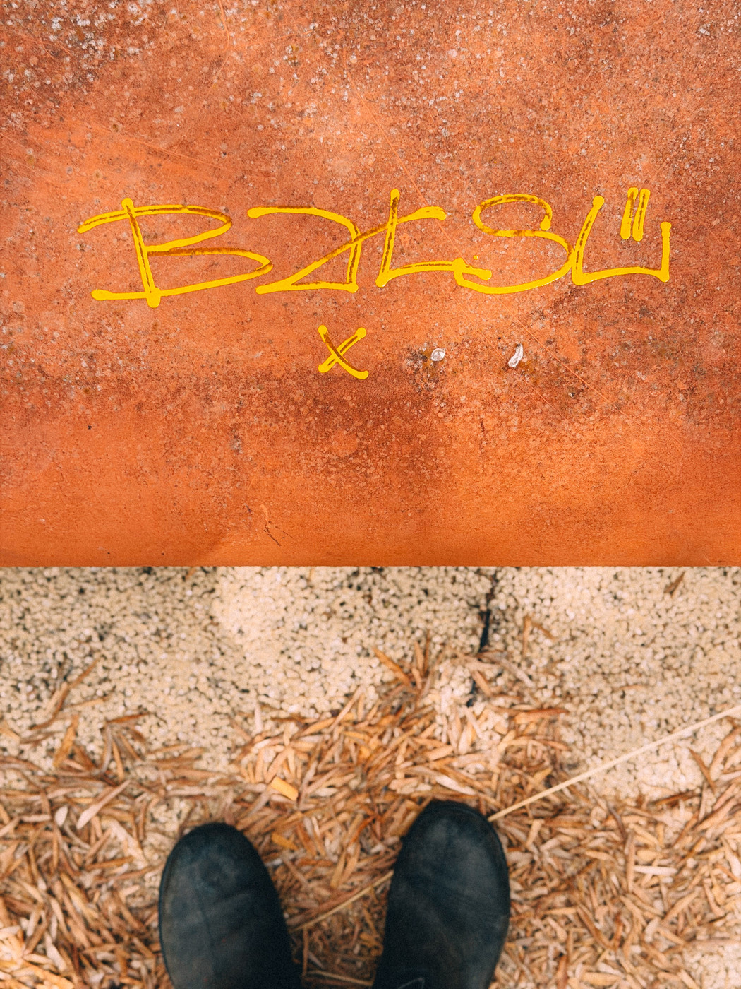 Yellow graffiti text on a rusty orange surface with a pair of black boots visible on wood chips and gravel below.