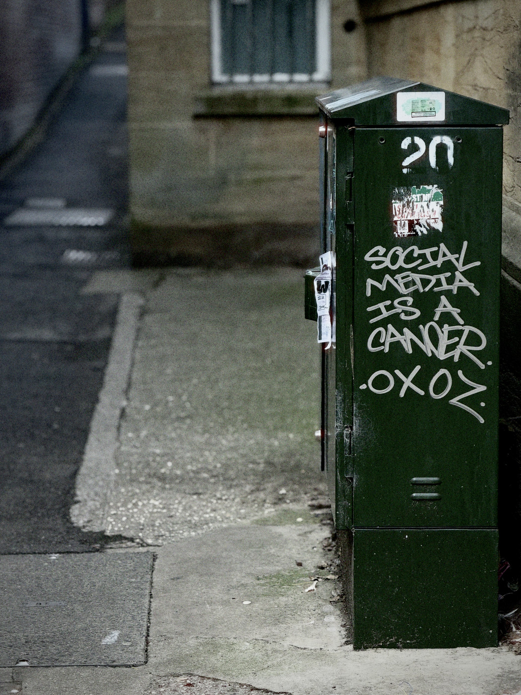 Green street utility cabinet tagged with graffiti reading “Social media is a cancer” and the signature “OXOZ”, quiet pavement behind.