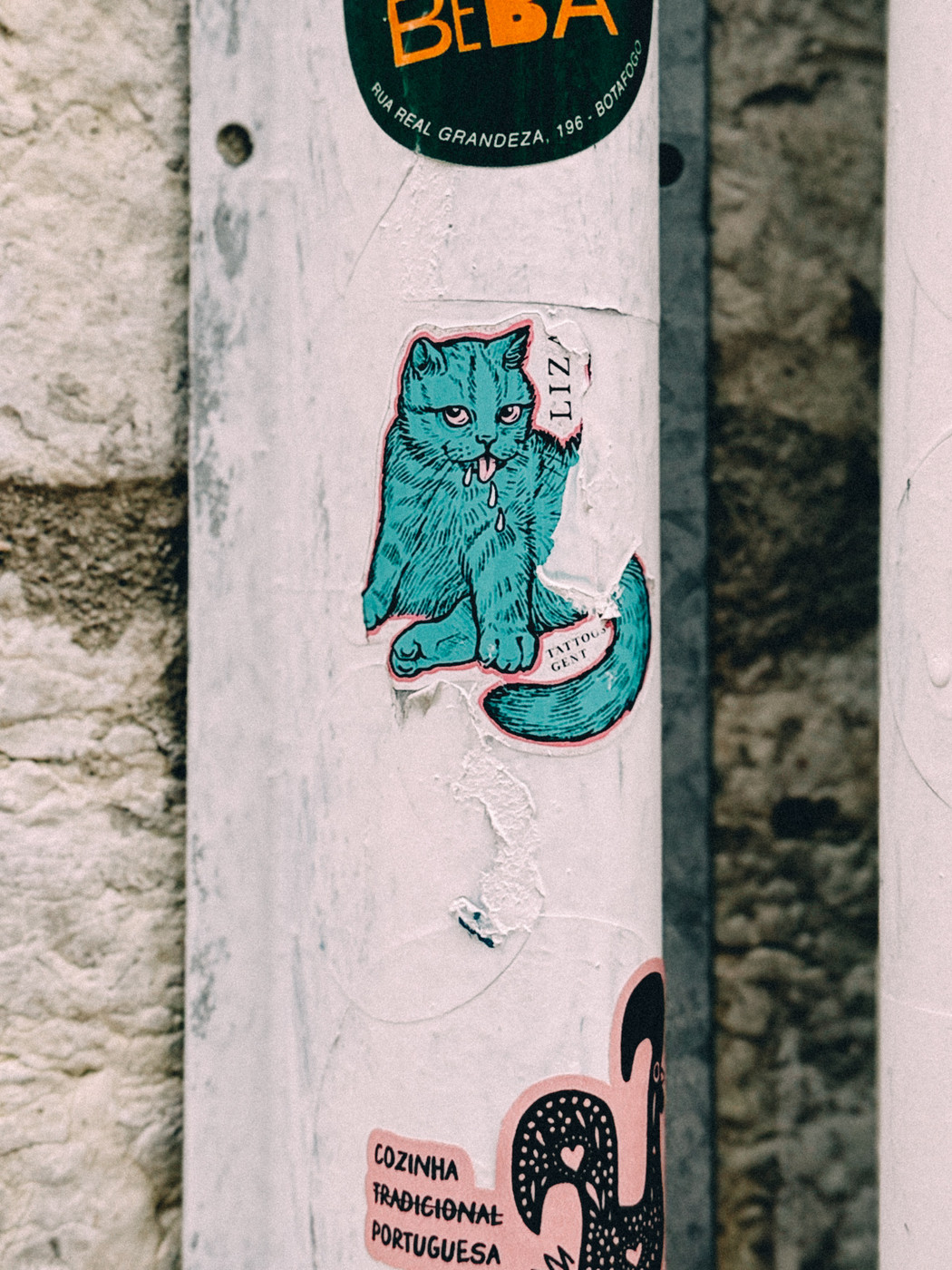 Colorful street art stickers on a white pole, featuring a blue drooling Cat, surrounded by other stickers including one with Portuguese text.…