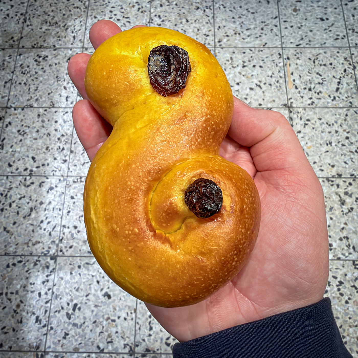 A hand holding a bright yellow saffron bun shaped like an S with both ends curled up and a raisin in each curl, above a speckled terrazzo floor.