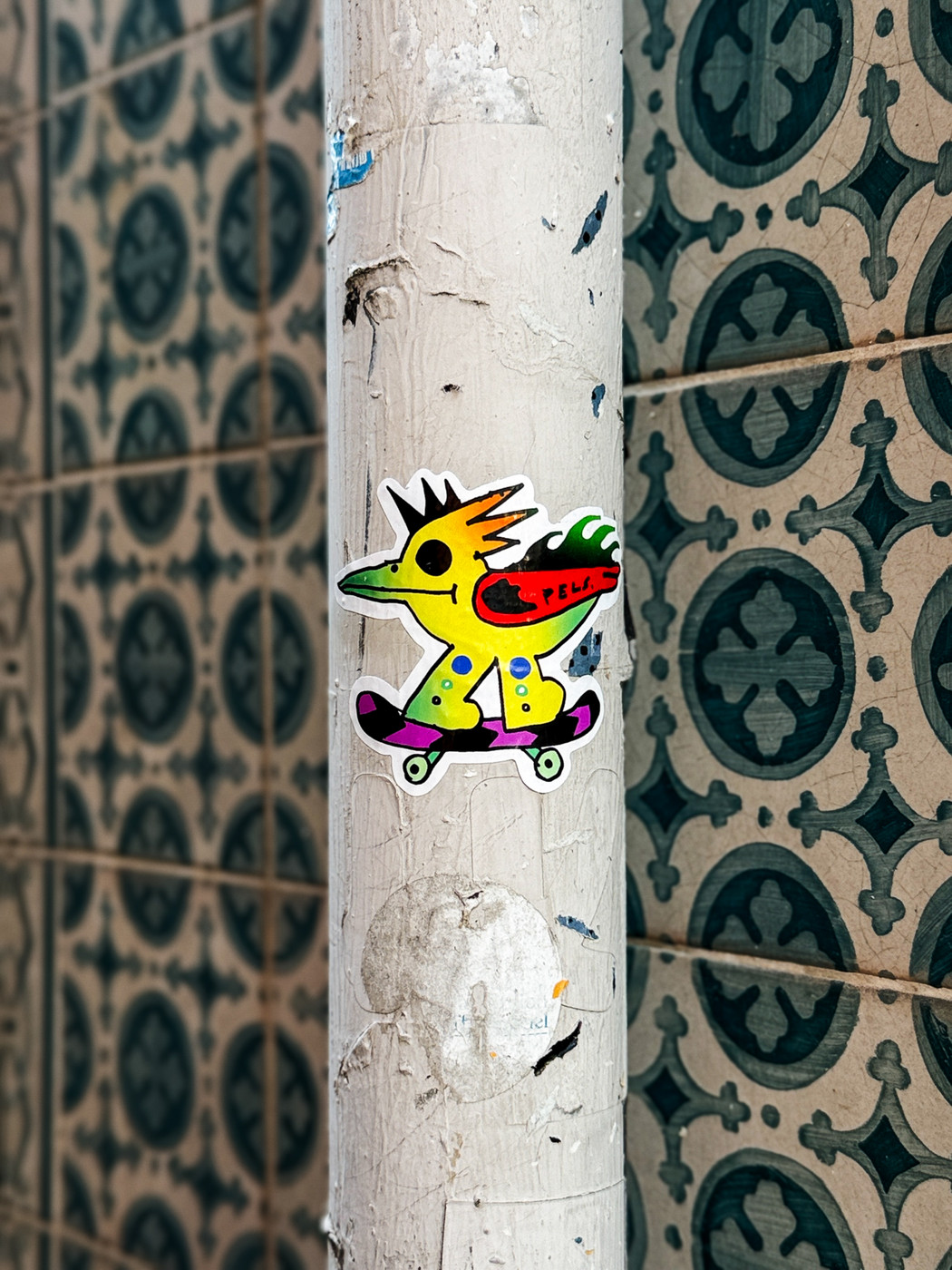 Colorful sticker of a cartoon bird with spiky hair riding a skateboard on a weathered white pole, with blue patterned tiles in the background.