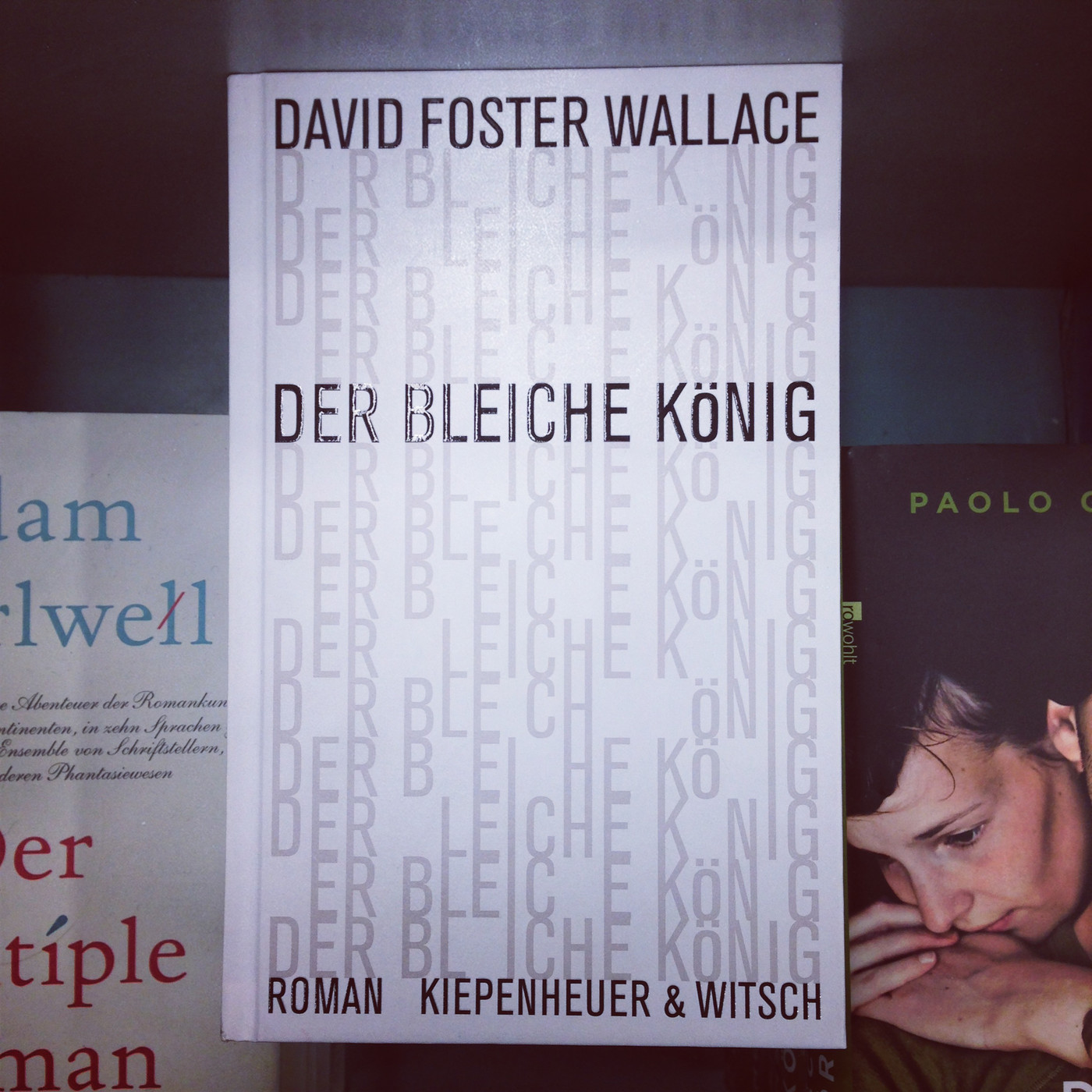 In einem leicht abgedunkelten Bücherregal steht die deutsche Ausgabe des Romans „Der bleiche König“ von David Foster Wallace (Kiepenheuer & Witsch) aufrecht im Zentrum des Bildes. Links davon ist der Anschnitt eines Buches mit hellem Cover und blauer sowie roter Schrift erkennbar. Rechts daneben steht ein dunkles Buch aus dem Rowohlt Verlag; darauf sind der partiell sichtbare Autorenname „PAOLO“ und das Gesicht einer nachdenklich nach unten blickenden Frau abgebildet. Das Foto hat eine sichtbare Vignettierung, wodurch die Ränder abgedunkelt wirken.
