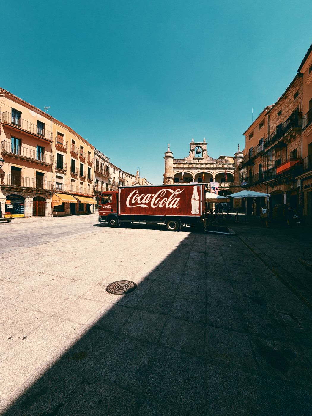 A red Coca-Cola delivery truck parked in a sunny European town square with historic buildings and a clock tower under a clear blue sky.