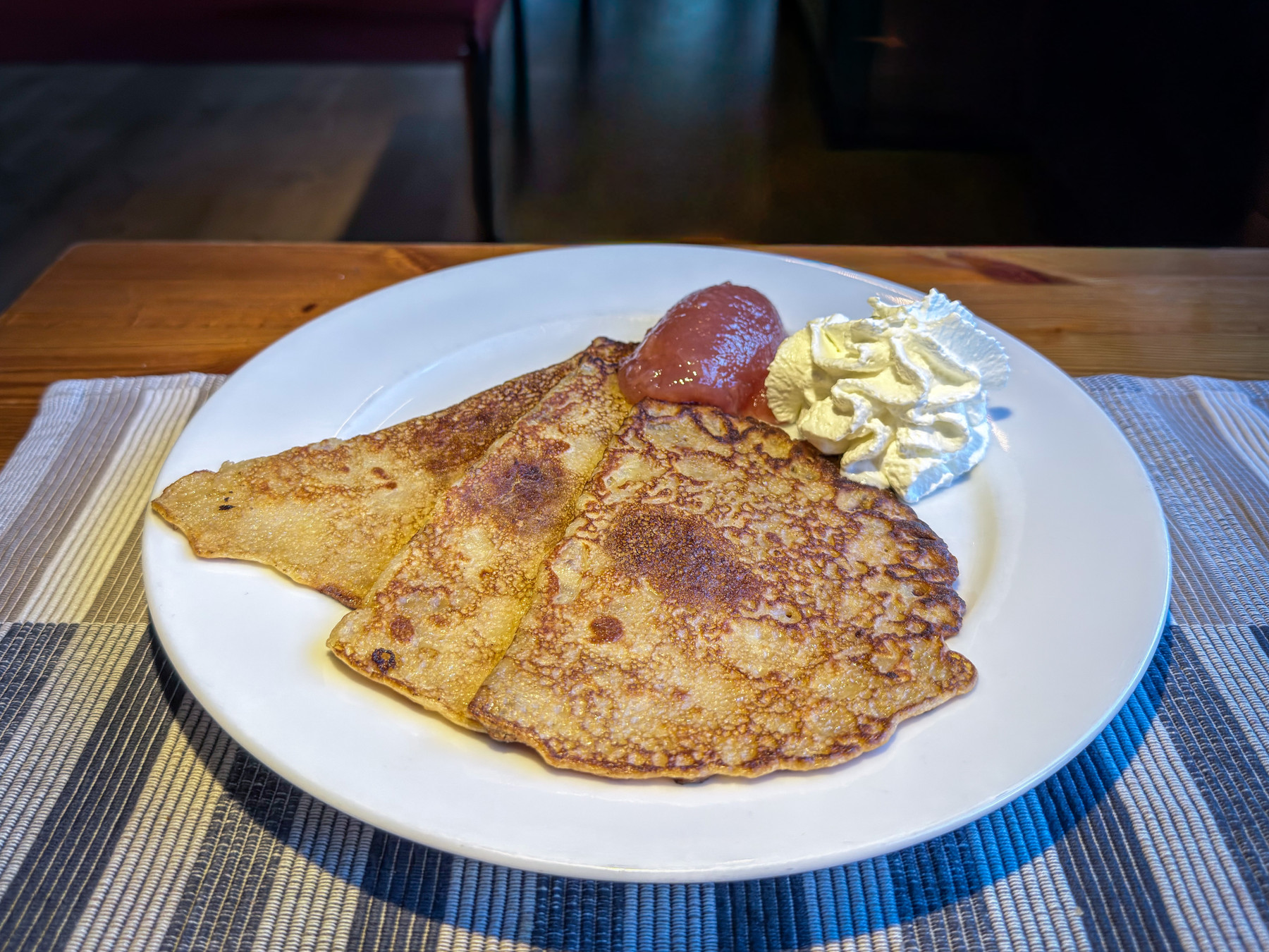 Three thin, golden Swedish pancakes folded into triangles on a white plate, served with a scoop of pink jam and a swirl of whipped cream, on a striped placemat.