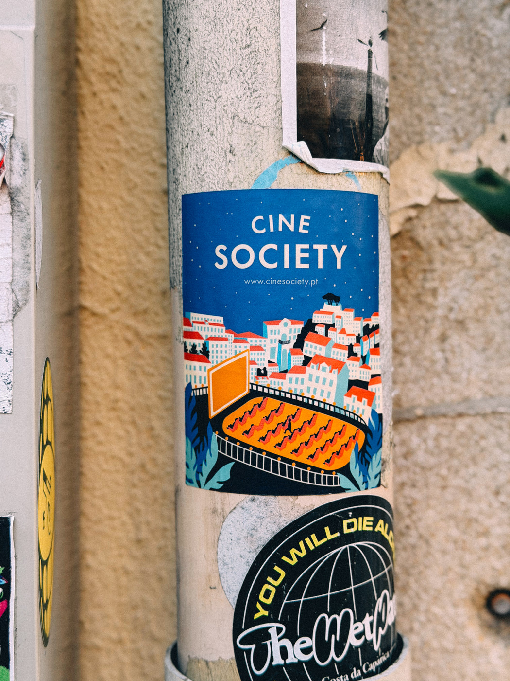 Colorful street sticker on a pole advertising “Cine Society” with an illustration of an outdoor cinema surrounded by vibrant city buildings, alongside other stickers and weathered textures.