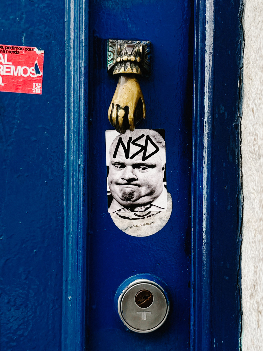 Close-up of a blue door with a brass hand-shaped door knocker, a circular lock, and a black-and-white sticker of a man’s face with “NSD” written above it.