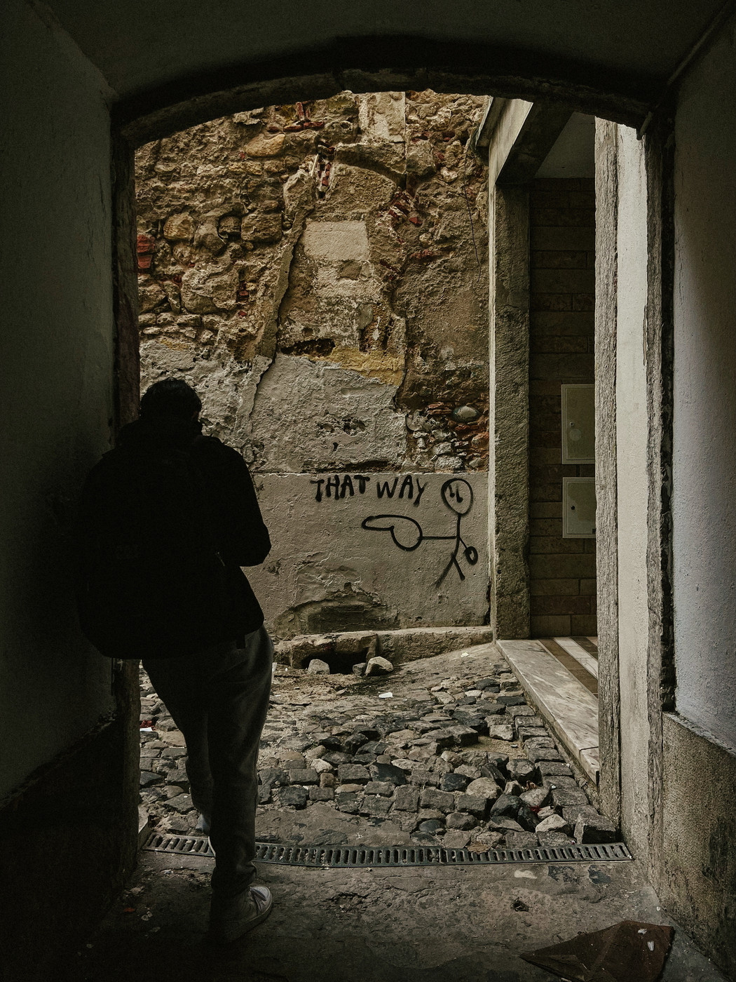 A person stands in a dim archway, looking at graffiti on a worn stone wall. The graffiti features a stick figure pointing with the words “THAT WAY” next to it. The ground is uneven with cobblestones.