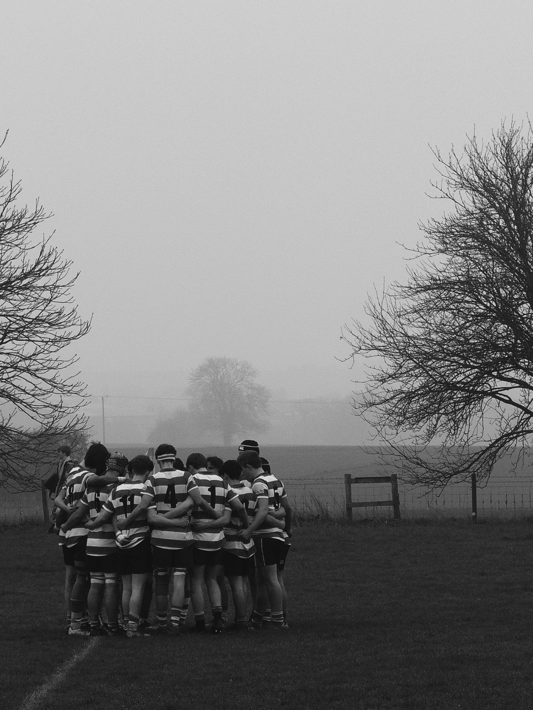 A black and white photo of a rugby team huddled together in a field. The players are wearing striped jerseys and shorts. The field is surrounded by trees and a fence. The sky is overcast and foggy. The mood of the image is calm and focused.
