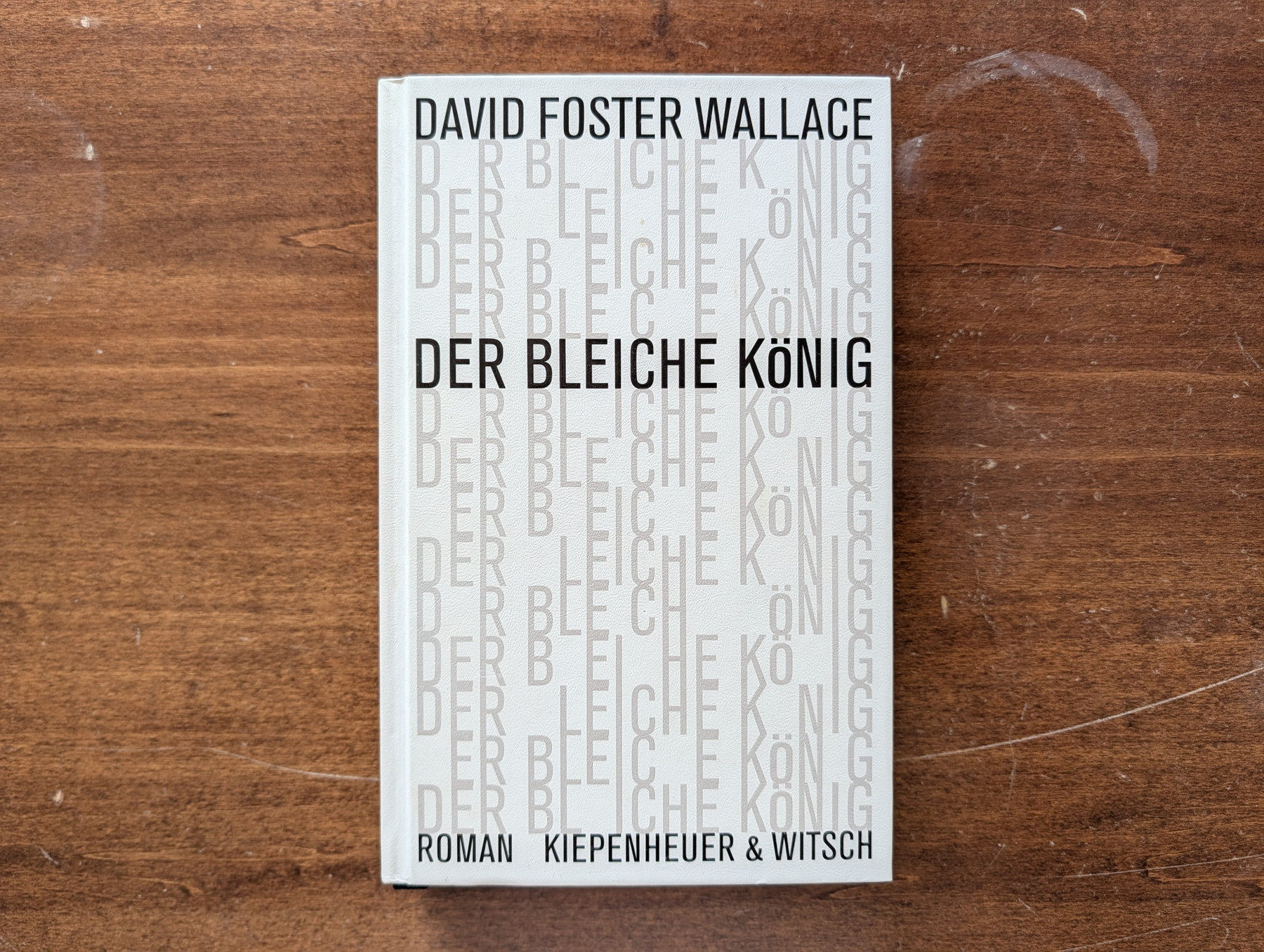 Auf einer braunen, leicht zerkratzten Holzoberfläche liegt die deutsche Ausgabe des Romans „Der bleiche König“ von David Foster Wallace. Das weiße Cover-Design besteht aus sich wiederholenden, hellgrauen Textzeilen des Titels, bei denen stellenweise Buchstaben fehlen. Im starken Kontrast dazu stehen der Autorenname oben, der Titel in der Mitte und die Verlagsangabe „ROMAN KIEPENHEUER & WITSCH“ unten in klarer, schwarzer und serifenloser Schrift.