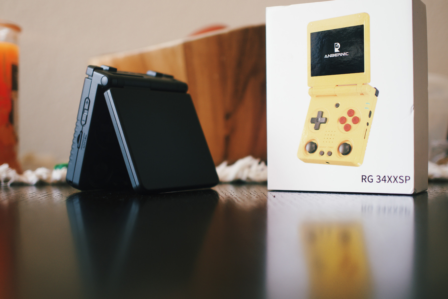 A black clamshell-style handheld gaming console sits partially open on a wooden surface beside its packaging box. The box shows a yellow version of the same device with the label “ANBERNIC RG34XXSP” printed on it. The background is softly blurred, giving a warm and cozy indoor atmosphere.