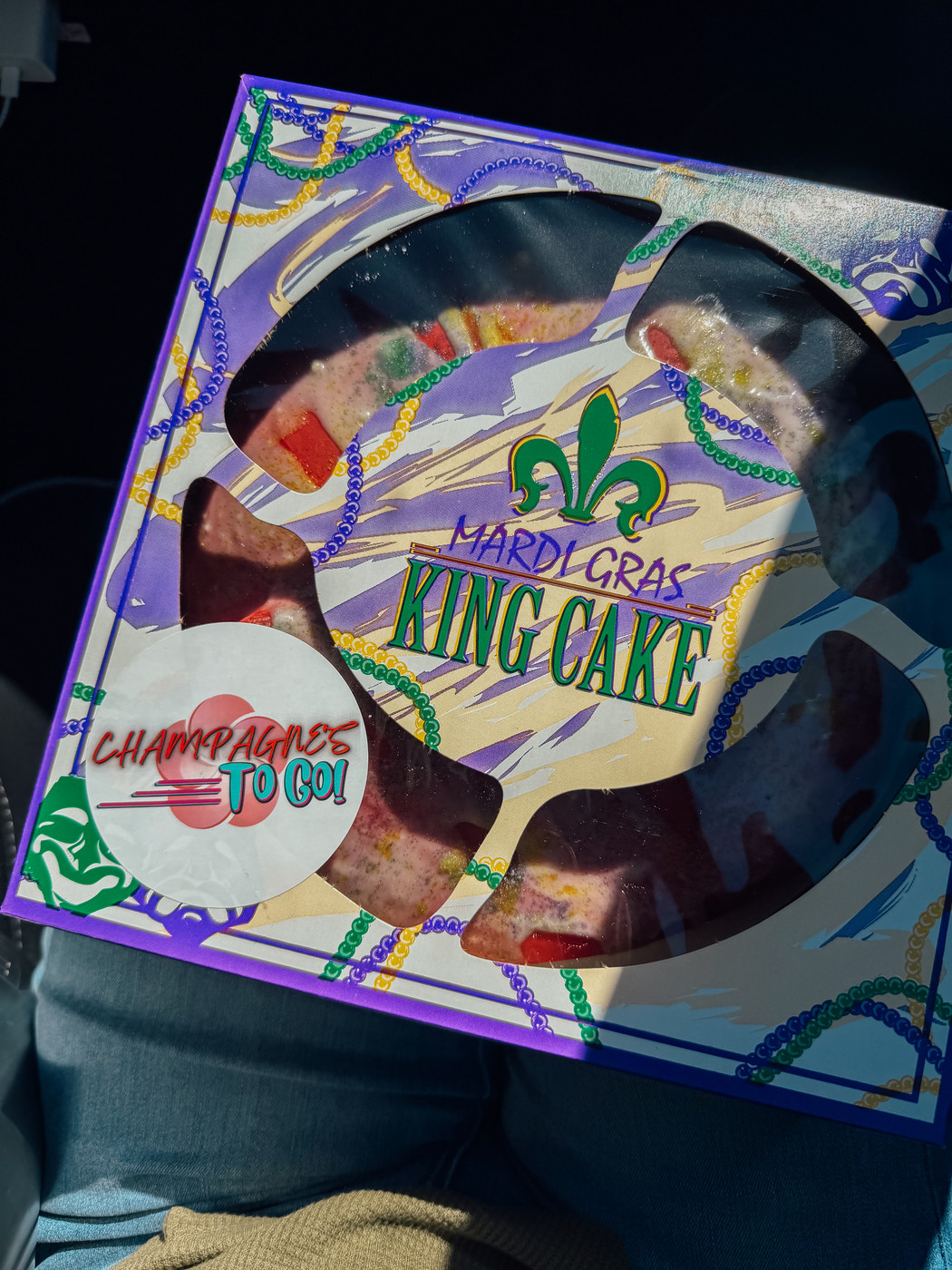 A colorful Mardi Gras King Cake is packaged in a festive box with a fleur-de-lis design.