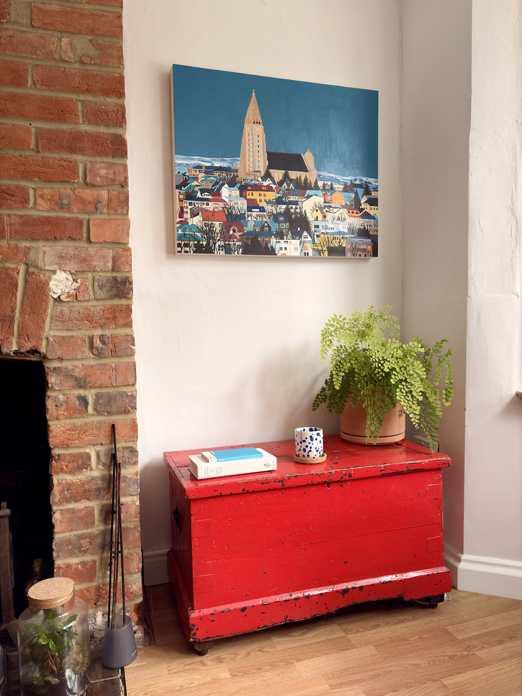 A bright red, distressed wooden chest sits beside a brick fireplace, topped with a potted fern, a spotted mug and books, beneath a colourful painting of a town and tall church against a blue sky.