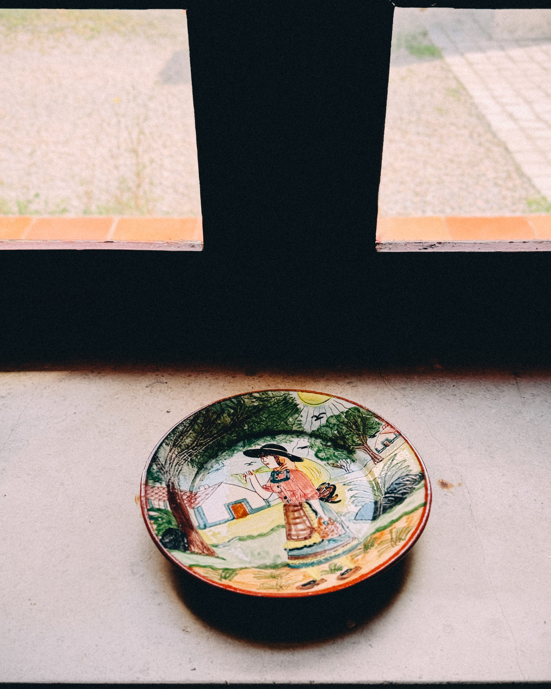 Colorful hand-painted ceramic plate depicting a woman in traditional clothing with trees, houses, and a sunlit countryside background, placed on a windowsill with natural light.