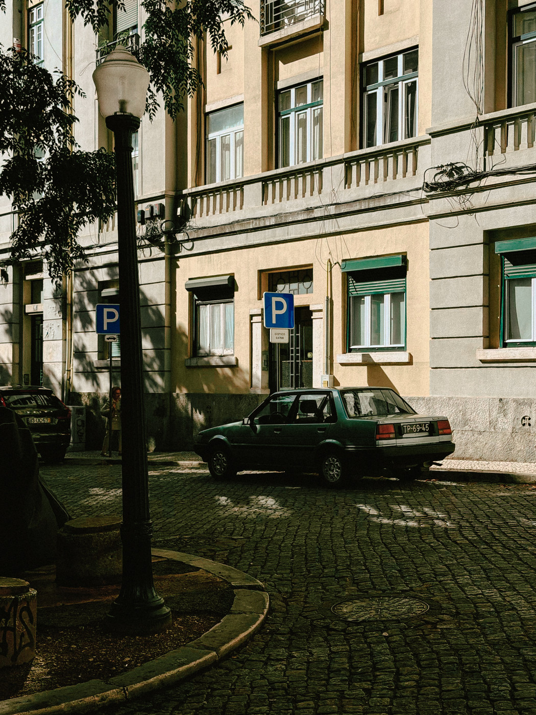 A dark green sedan is parked on a cobblestone street in front of a beige apartment building with green-trimmed windows, a lamppost, and parking signs under partial tree shade.
