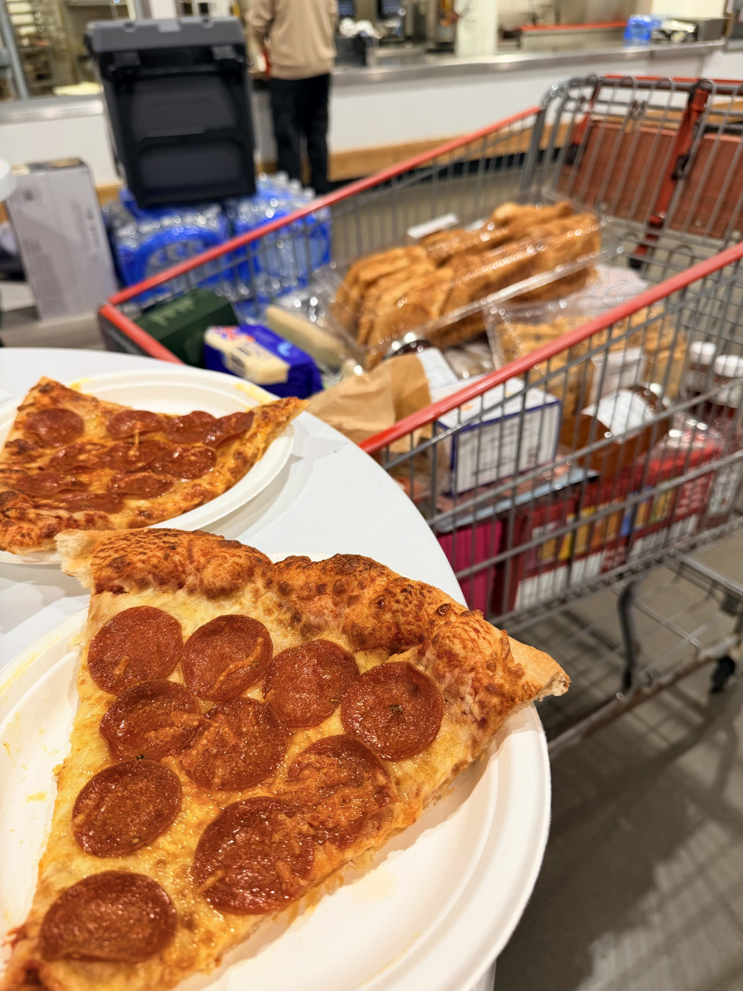 Two large slices of pepperoni pizza sit on white disposable plates in the foreground, photographed at a warehouse food court. A shopping trolley filled with bulk groceries, bottled water, and bakery items stands beside the table, while a customer waits at the service counter in the background.