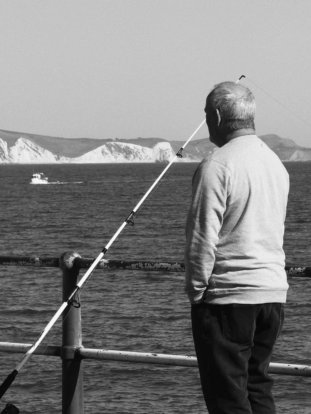 A black and white photo of an older man seen from behind fishing off a seaside railing, with cliffs and sea in the background.