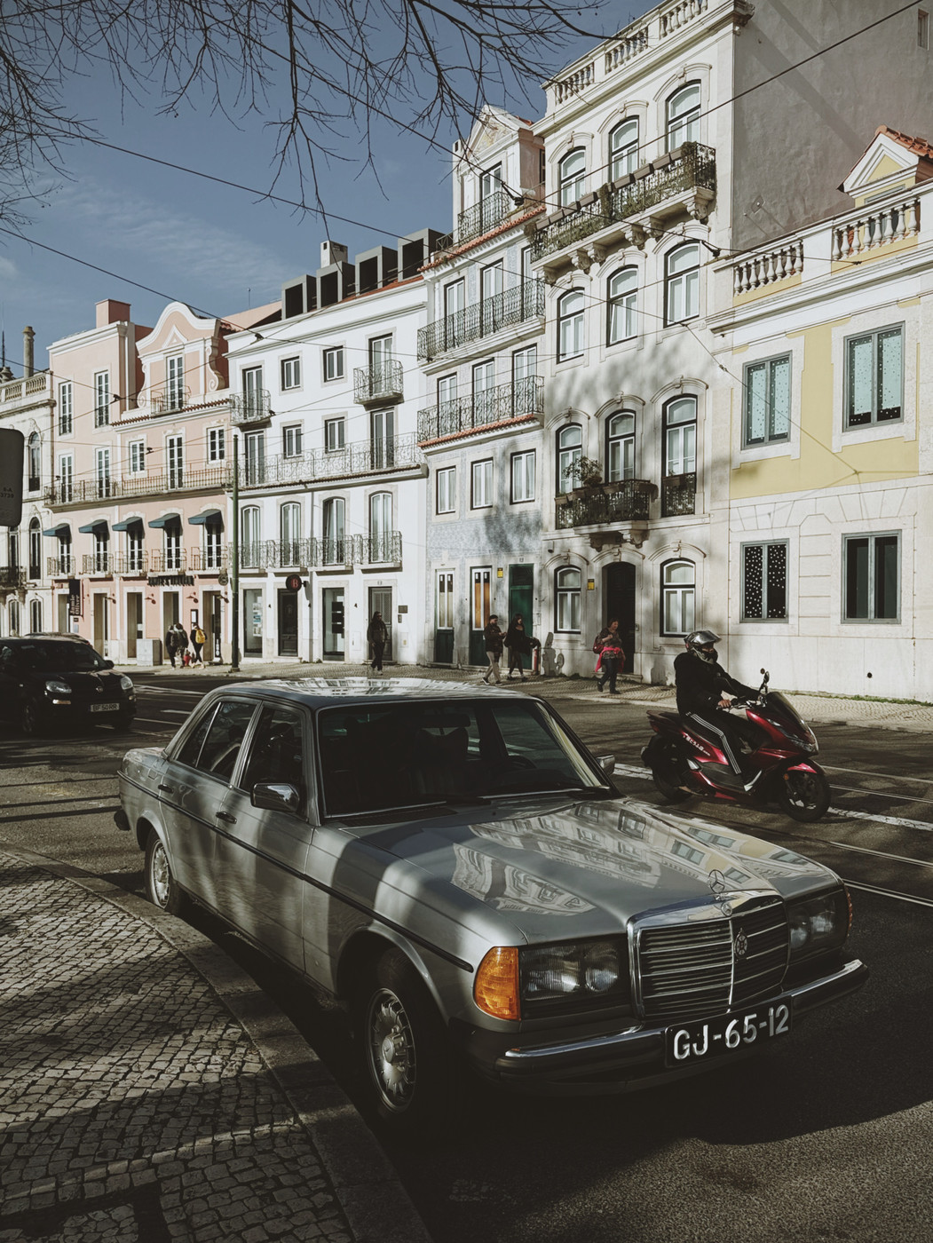 A vintage silver Mercedes-Benz is parked on a cobblestone street in front of pastel-colored European buildings, with a motorcyclist riding by and shadows of bare tree branches cast on the scene.