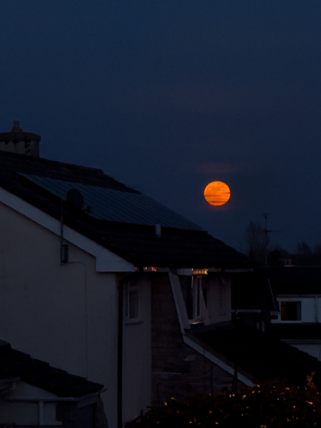 A large orange sun sets behind the rooftops of suburban houses. The sky is a deep blue, and the houses are silhouetted against it. The mood is calm and peaceful, suggesting the end of a day.