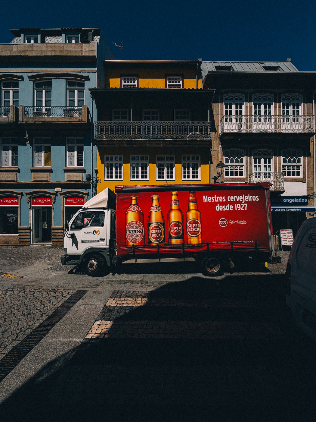 A delivery truck with a red Super Bock beer advertisement is parked on a cobblestone street in front of colorful European buildings under a clear blue sky.