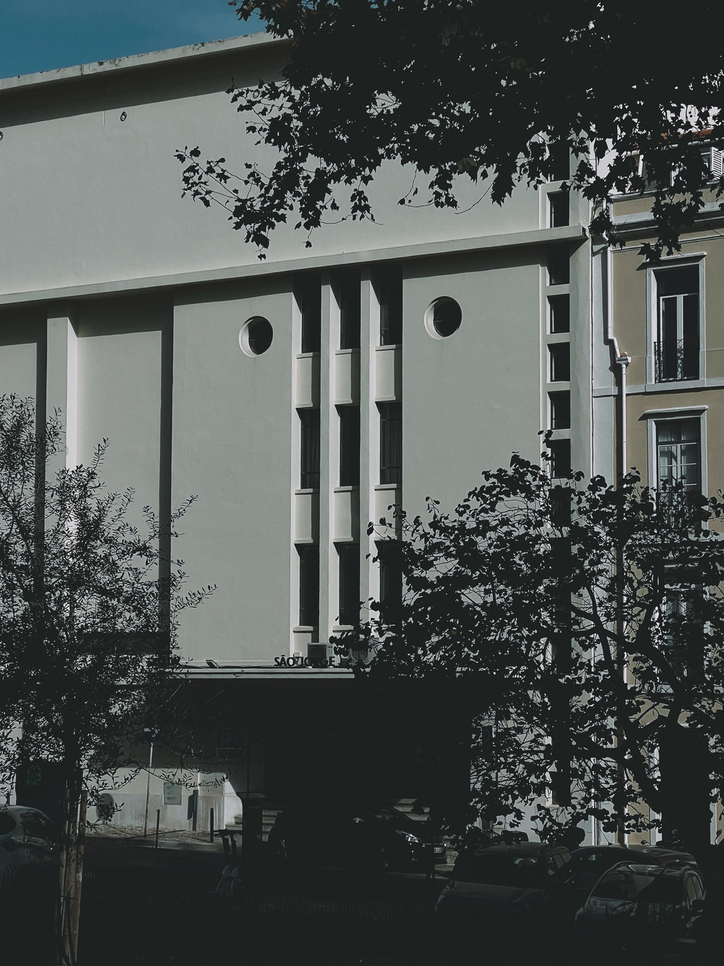 An angular white building with vertical windows and circular details, partially shaded by tree branches, with parked cars and street elements in the foreground.