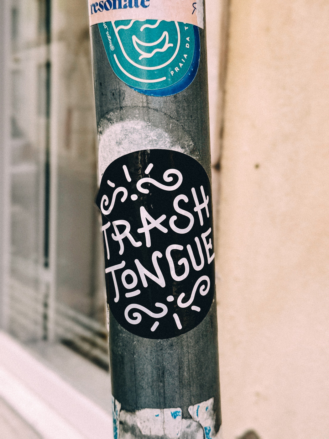 A black circular sticker with the white handwritten words “TRASH TONGUE” and decorative lines is attached to a weathered metal pole on a city street.