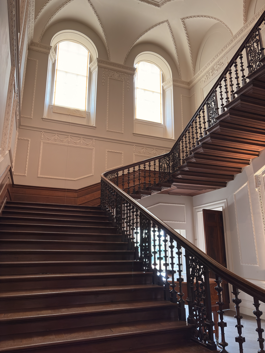 A grand wooden staircase with ornate dark balustrades rises to a landing beneath a high vaulted ceiling decorated with elegant plaster mouldings. Two tall arched windows flood the space with soft daylight, highlighting the pale walls, carved architectural details and polished timber steps.