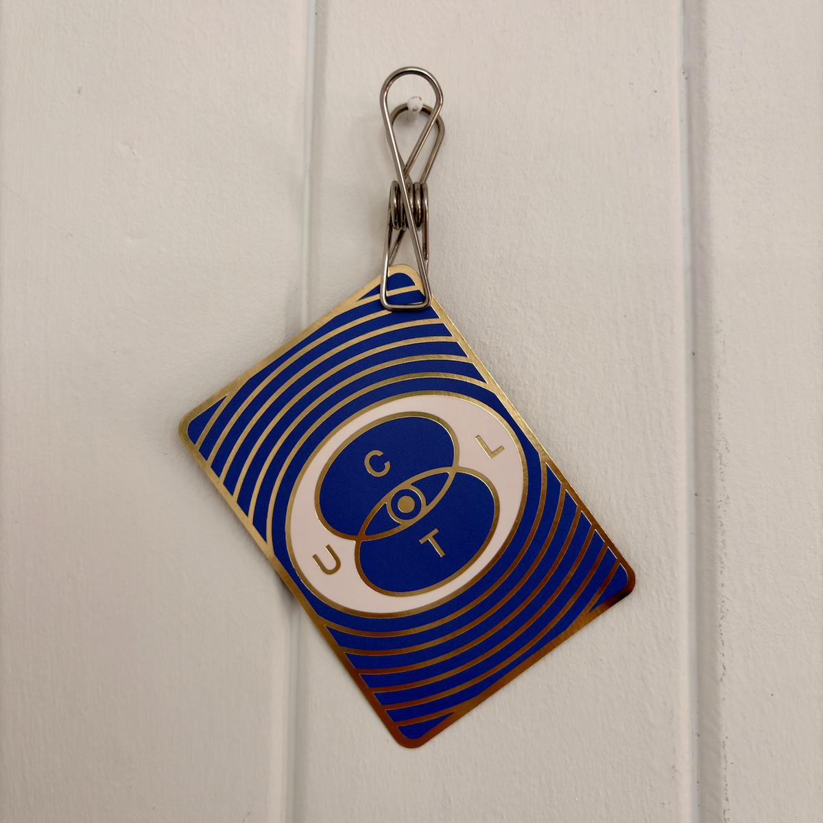 A blue and gold patterned card hangs from a metal clip on a white panelled wall.