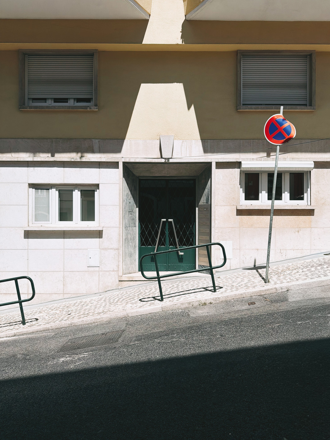 A slanted street with a beige building, green door, and barred windows, featuring a bent no-parking sign and metal railings along the sidewalk.