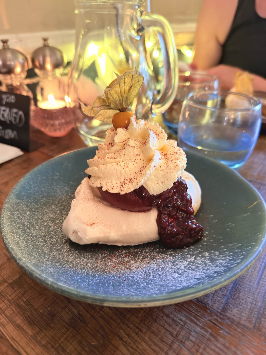 Meringue dessert topped with piped cream, dark berry compote, and a physalis fruit, on a teal plate at a candlelit table.