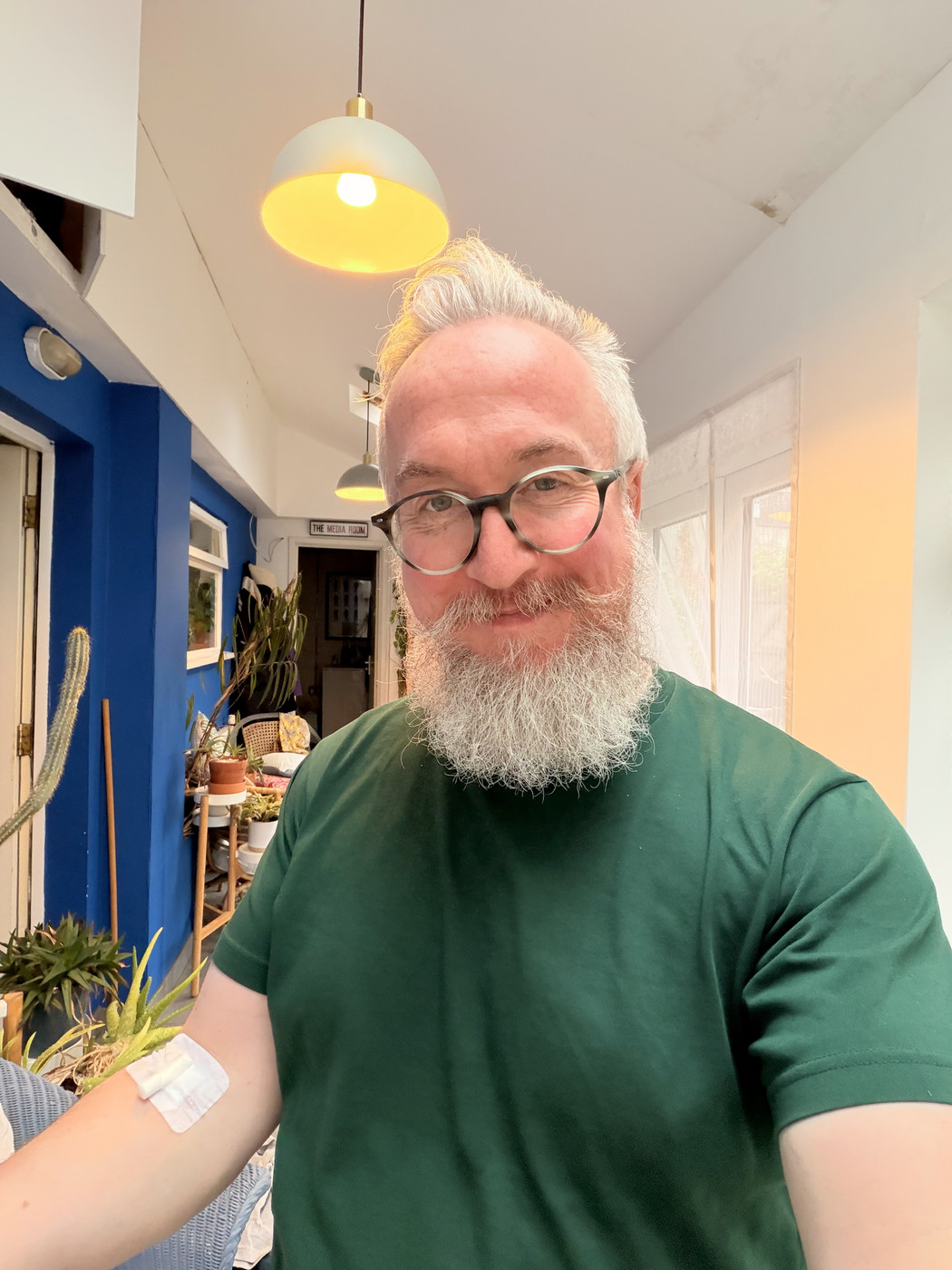 A man with a white beard and glasses, wearing a green t-shirt, with a white bandage on his inner elbow, likely after donating blood.