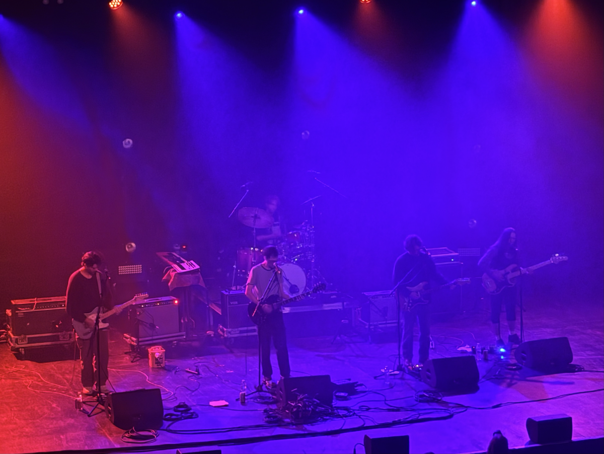 A five-piece band performs on stage under a mix of orange and blue lighting. The setup includes a drummer at the back, two guitarists, a bassist, and a keyboard player. Cables and equipment are spread across the stage floor, and the scene is softly illuminated by stage spotlights through a light haze.