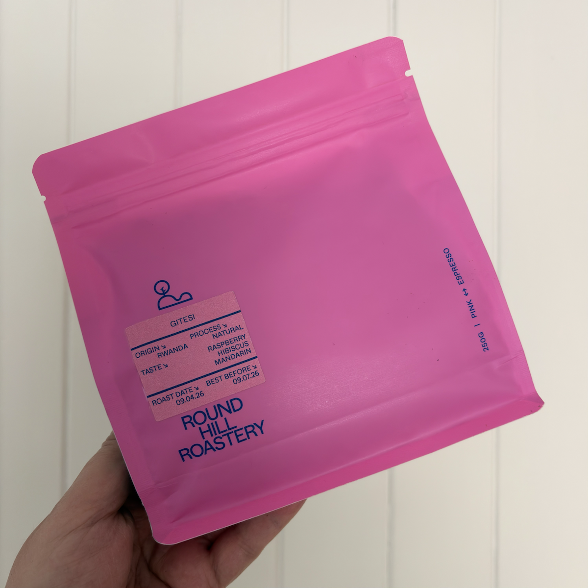 Hand holding a bright pink coffee bag from Round Hill Roastery against a white panelled background. The label identifies the coffee as Gitesi from Rwanda, with tasting notes of raspberry, hibiscus and mandarin.