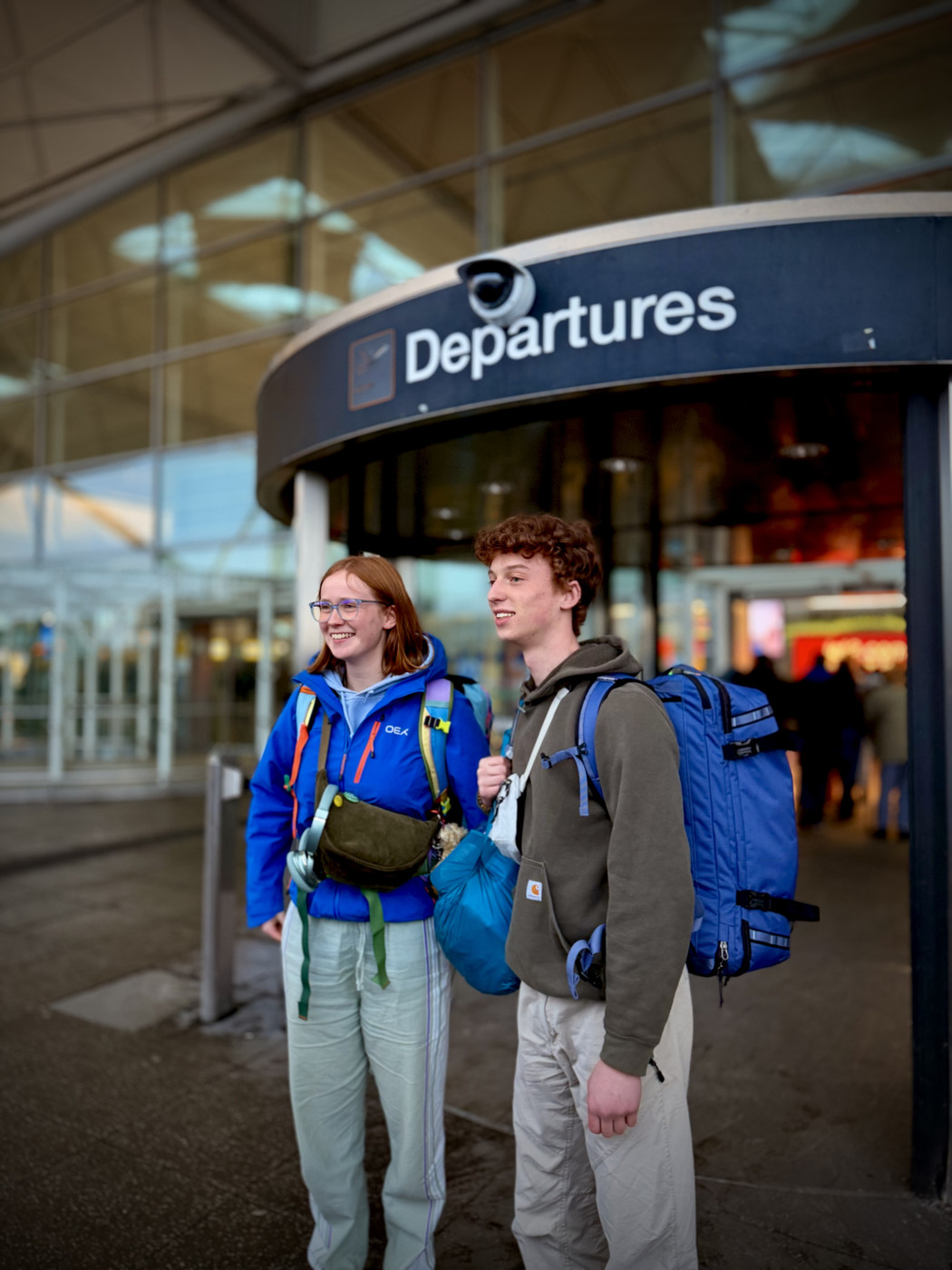 two young people standing outside an airport terminal beneath a large “Departures” sign. Both are wearing outdoor clothing and carrying large backpacks, suggesting they are about to travel or go on a longer trip.

They are standing side by side, looking off to the left, appearing relaxed and expectant. Behind them is a modern glass-fronted airport building, with other travellers and the entrance area slightly out of focus in the background. The overall mood feels calm and anticipatory, like the moment just before beginning a journey.