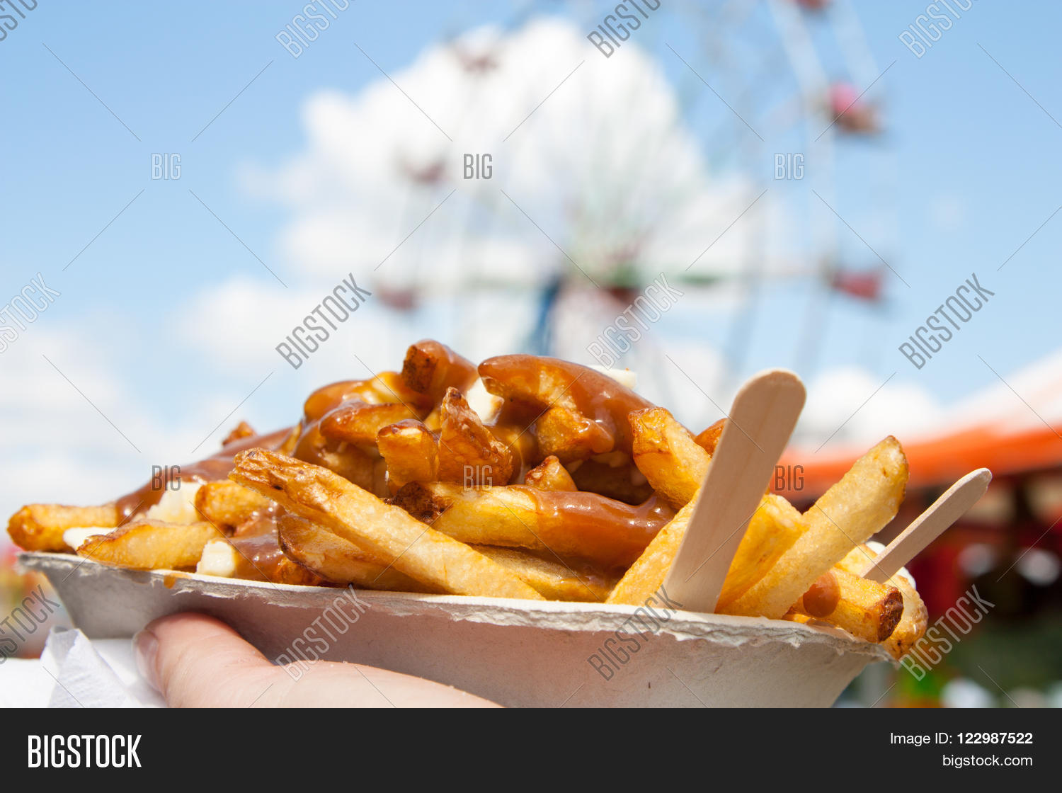A stock photo of a hand holding a paper tray of poutine — French fries topped with gravy and cheese curds with two wooden forks sticking out — held up against a blue sky with a Ferris wheel blurred in the background.