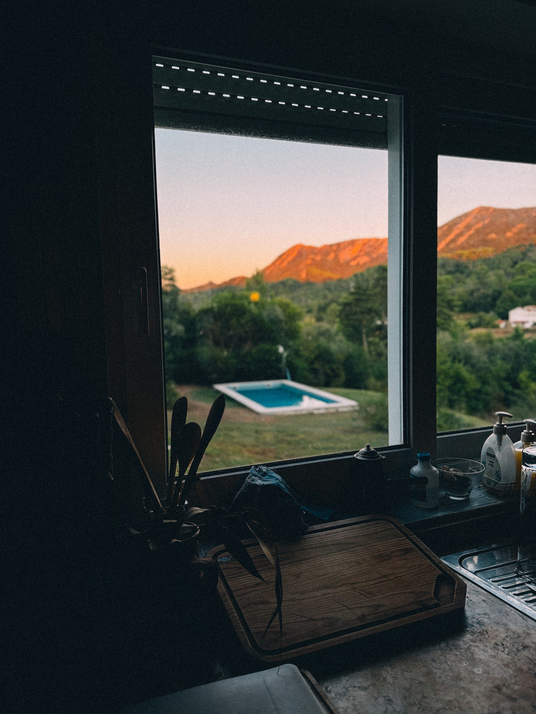 View from a dimly lit kitchen window overlooking a backyard with a pool, surrounded by greenery and mountains illuminated by the warm light of sunset.