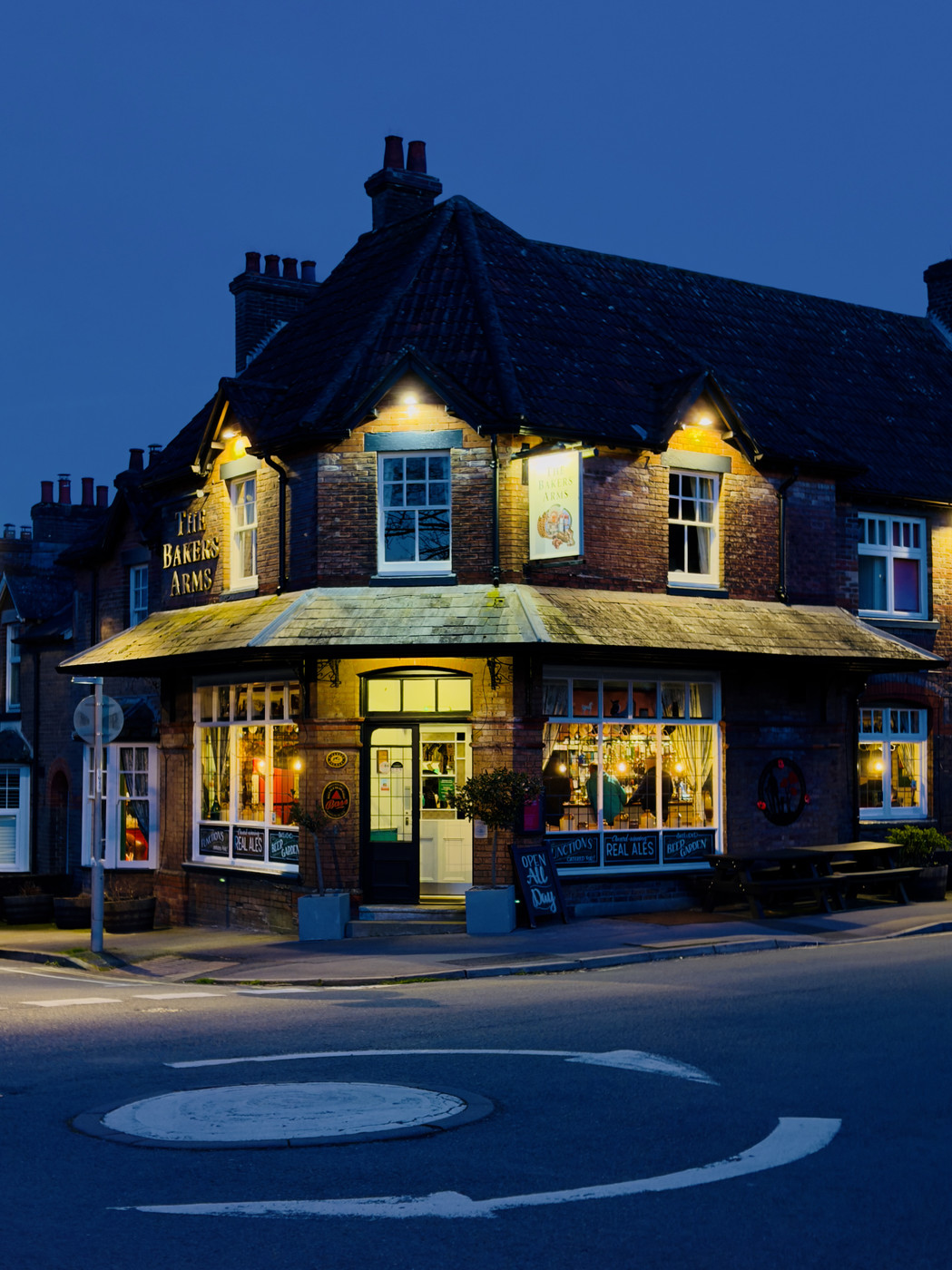 A brick building with a green roof and lit windows stands at a street corner. The building is a pub called "The Bakers Arms," with warm lights glowing from inside, creating a cozy and inviting atmosphere. Potted plants flank the entrance, and a sign advertises "Real Ale Daily." The scene is set during twilight, with a deep blue sky overhead, suggesting a peaceful evening in a quaint village or small town.