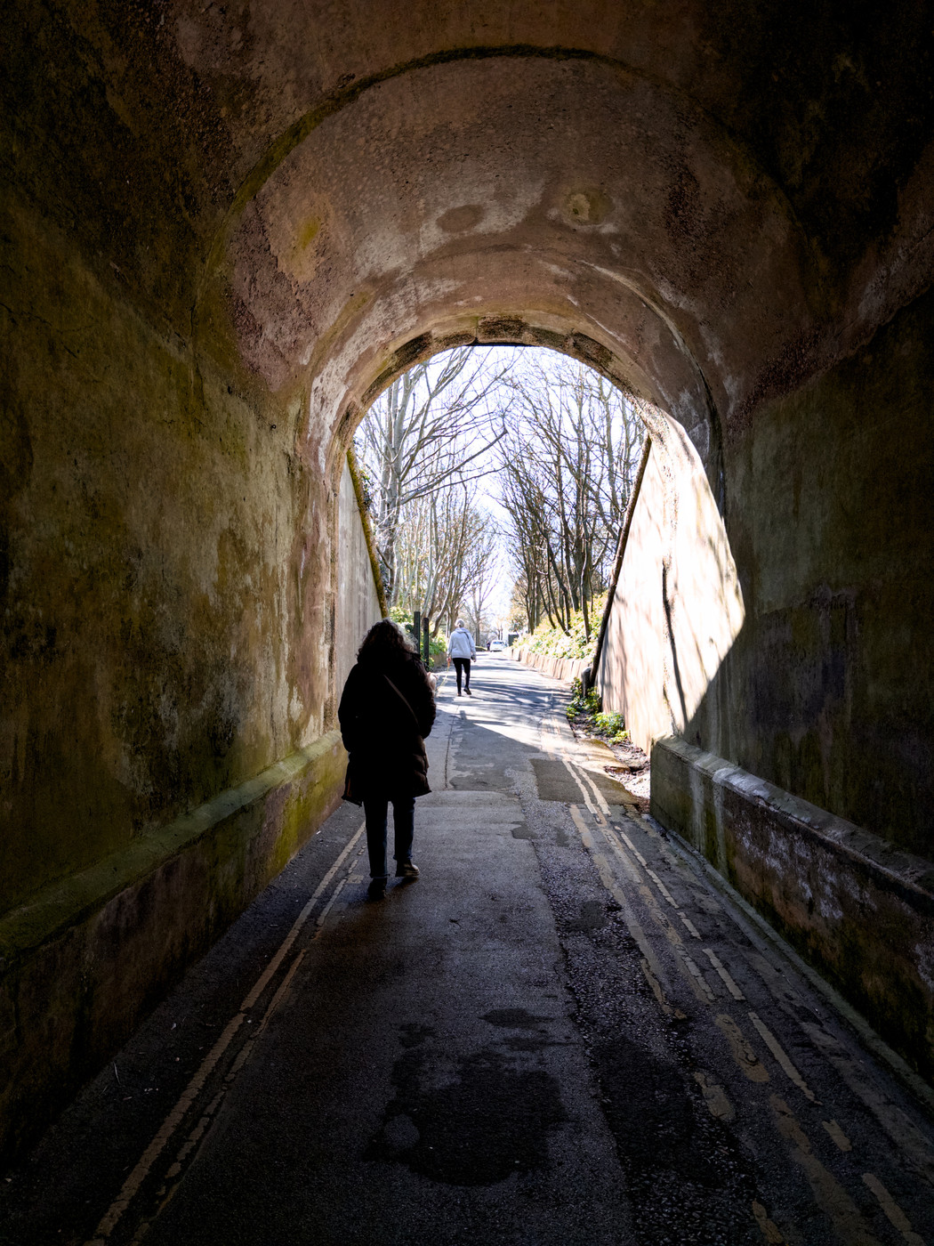 A person in a dark coat walks through a dimly lit tunnel towards a bright, tree-lined road. The tunnel walls are aged and mossy, creating a moody, atmospheric scene. The contrast between the dark tunnel and the sunlit exit evokes a sense of transition and hope.