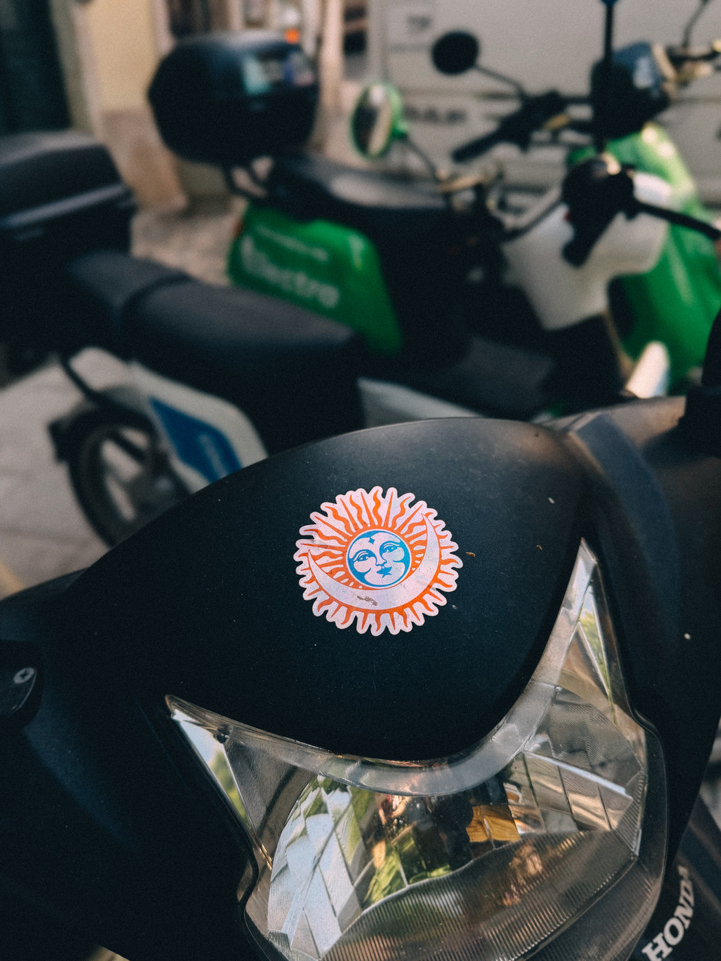 Close-up of a black scooter with a colorful sun and moon sticker on its front, parked among other green and white scooters on a street.