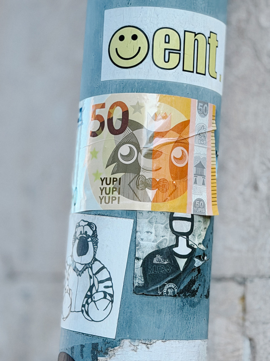 A blue pole covered with various stickers, including a colorful fake 50 Yupi banknote with cartoonish eyes, and a simple drawing of a striped animal resembling a tiger.