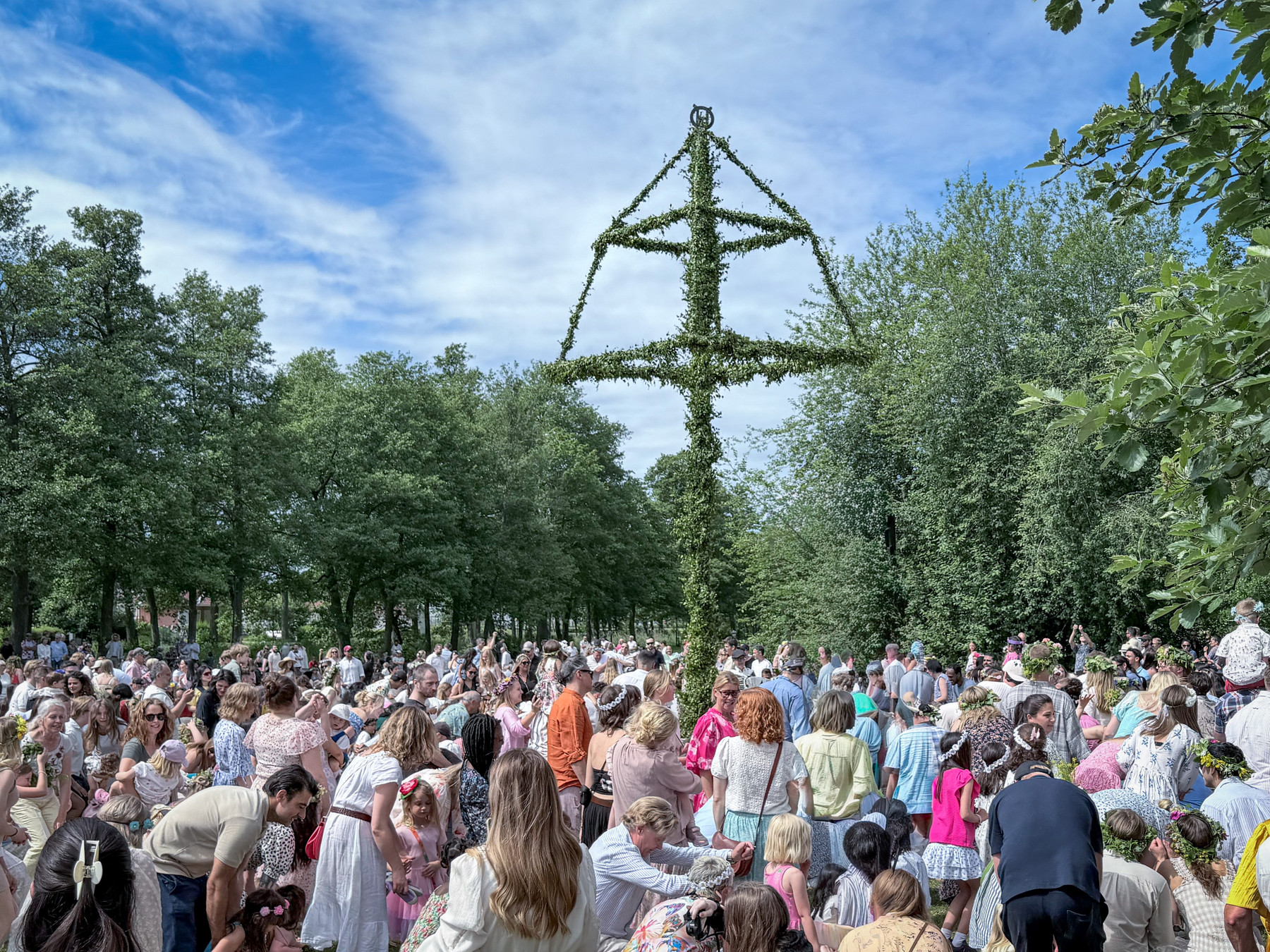 A tall maypole covered in green leaves and flowers rising above a large crowd gathered on the grass, under a blue sky with scattered clouds.