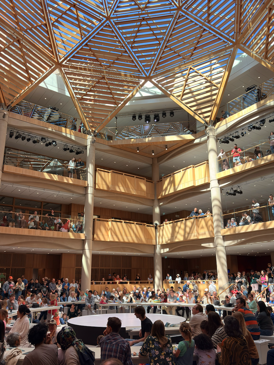Large indoor atrium with a geometric wooden lattice roof, where crowds gather around a circular central stage and watch from multiple balcony levels above.