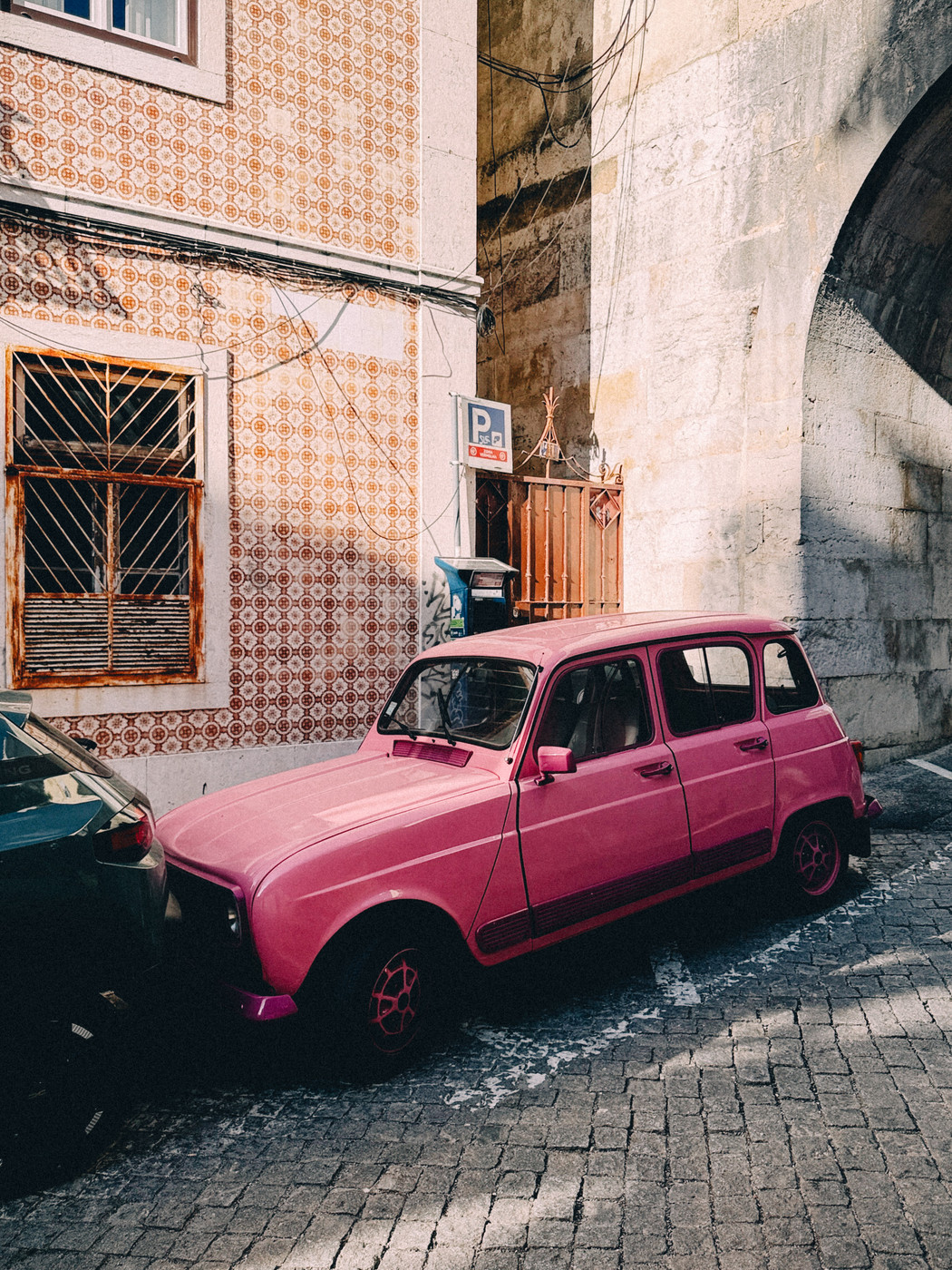 Bright pink vintage car parked on a cobblestone street next to a tiled building and stone archway in a sunny urban setting.