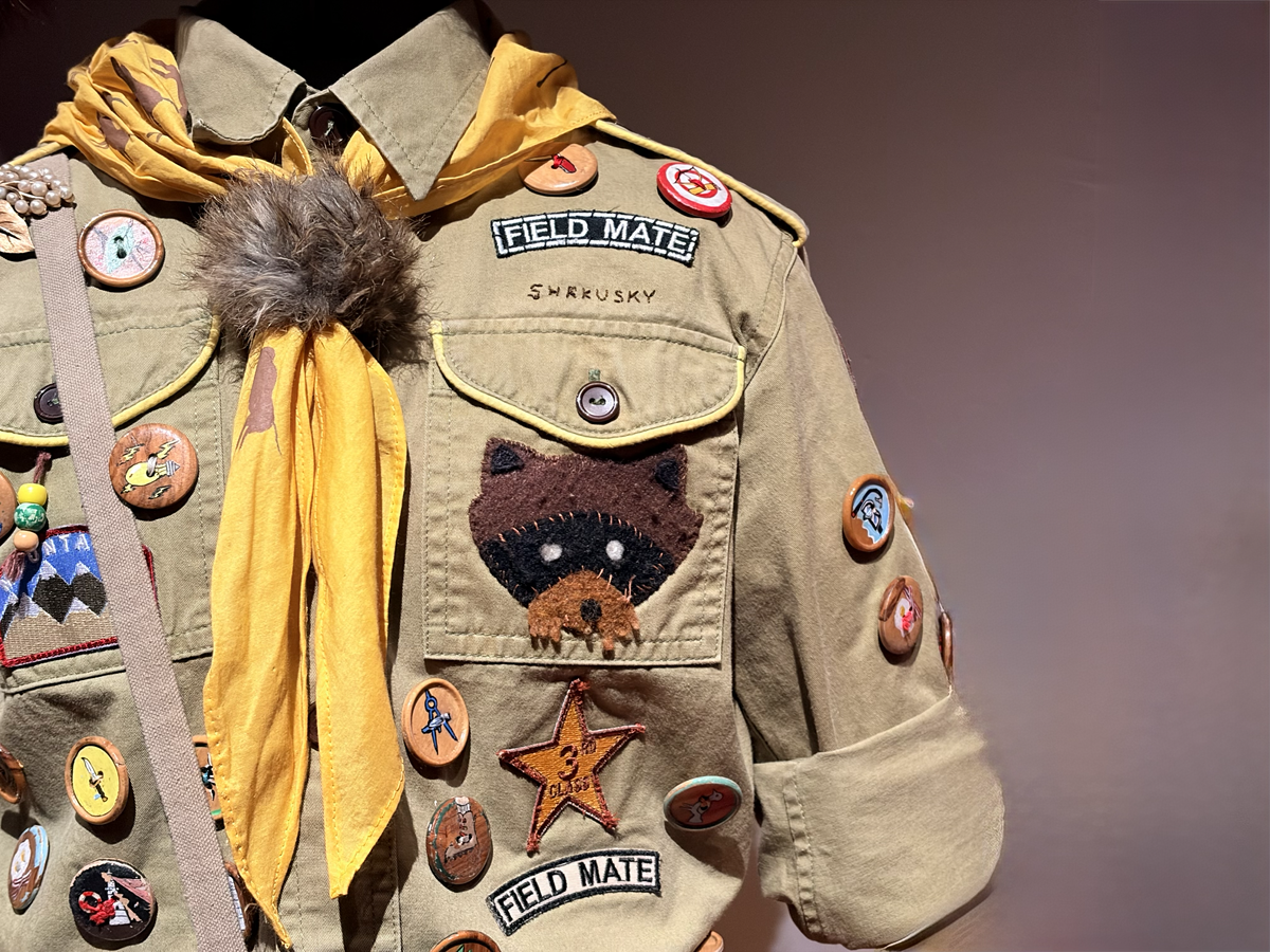 Close-up of a khaki scout-style jacket decorated with numerous badges and patches, including a stitched animal face, star emblem and ‘Field Mate’ labels, with a yellow neckerchief tied at the collar.