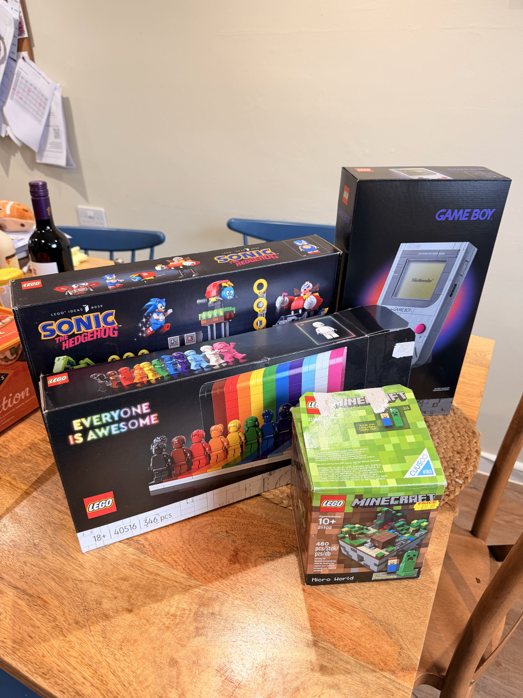 A wooden table holds four LEGO sets, including Sonic the Hedgehog, Game Boy, and Minecraft. The sets are colourful and new, suggesting excitement and anticipation. The background shows a beige wall and blue chairs, creating a cosy and inviting atmosphere.