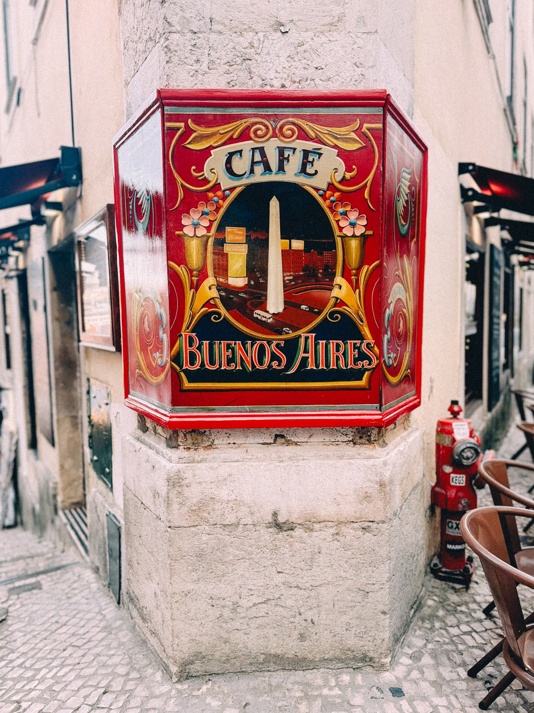 Colorful street sign for “Café Buenos Aires” with decorative flowers and an image of the Buenos Aires Obelisk, mounted on a stone corner wall along a narrow cobblestone street with outdoor seating.