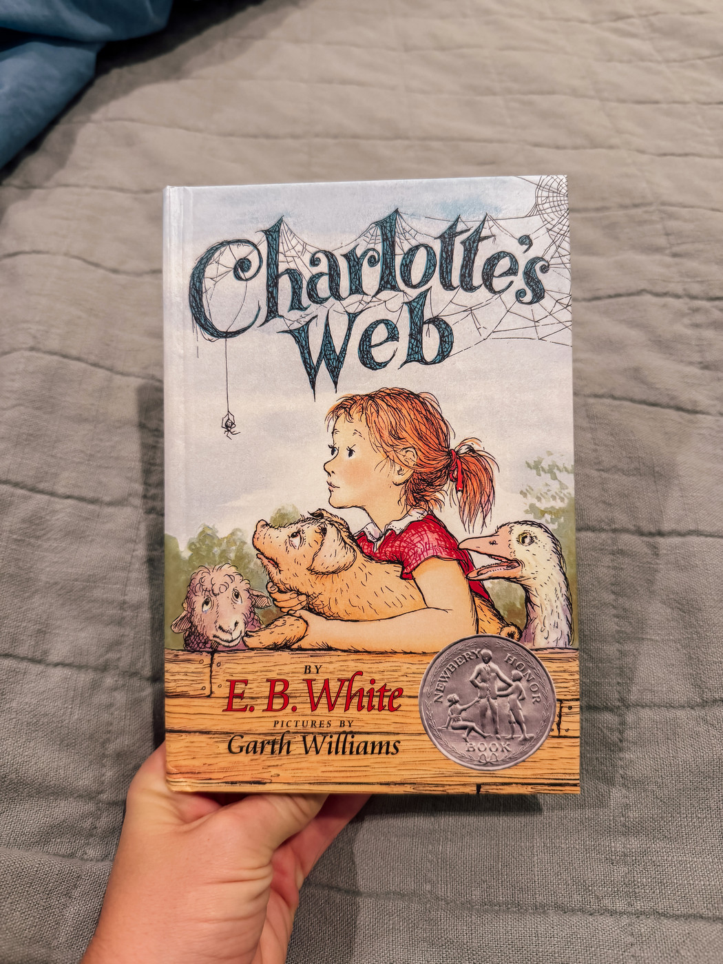 Auto-generated description: A hand holds a copy of Charlotte’s Web by E.B. White, featuring an illustrated cover of a girl leaning on a wooden fence with animals, accompanied by a Newbery Honor badge.