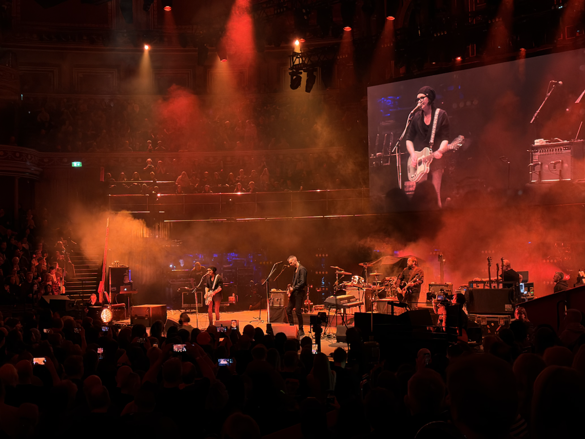 A live band performs on a large stage under red lighting and smoke effects, with a crowd watching and a large screen showing a close-up of the lead singer playing guitar.