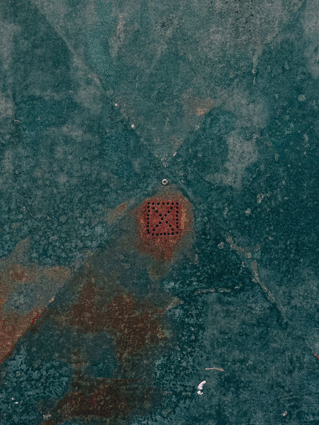 Weathered teal metal surface with a small rusted square vent featuring circular holes in the center.